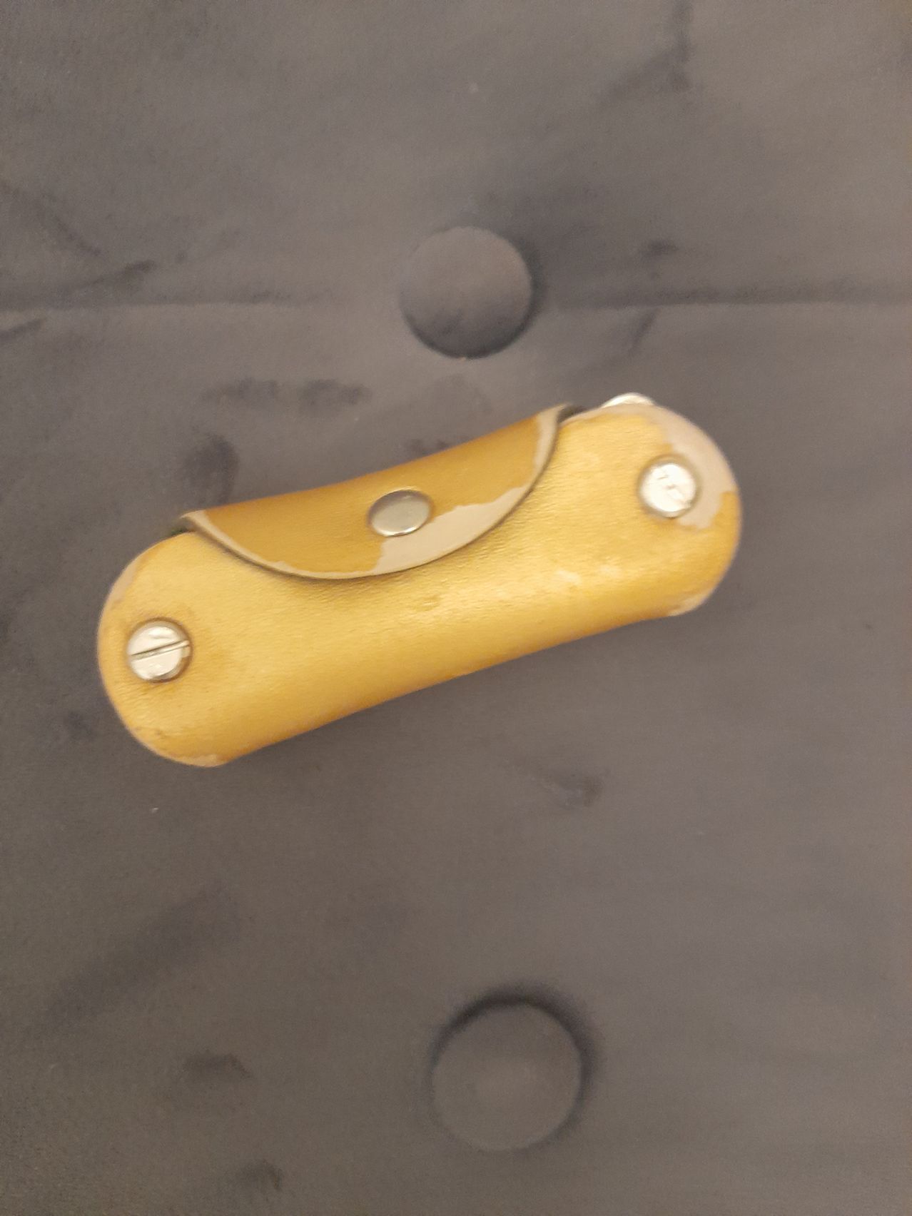 Ein Nahaufnahme eines kleinen, gelben, zylindrischen Objekts mit lederartigem Material und drei silbernen Schrauben auf einem grauen Hintergrund.