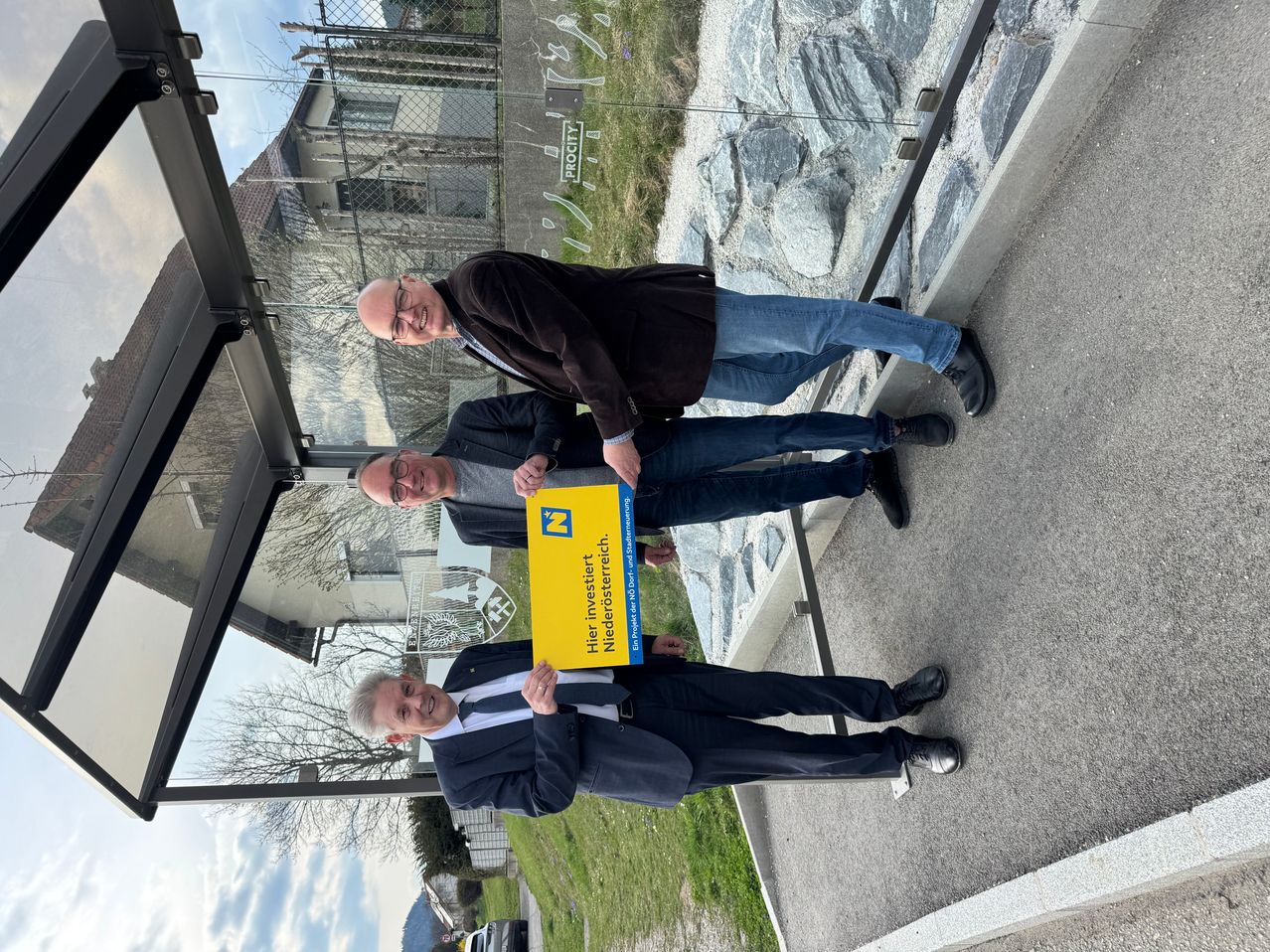Three men stand outside, holding a yellow sign that reads 'Here, invests Niederösterreich'. They are smiling and appear to be posing for a photo. Behind them, there is a fence with a rock wall and a building.
