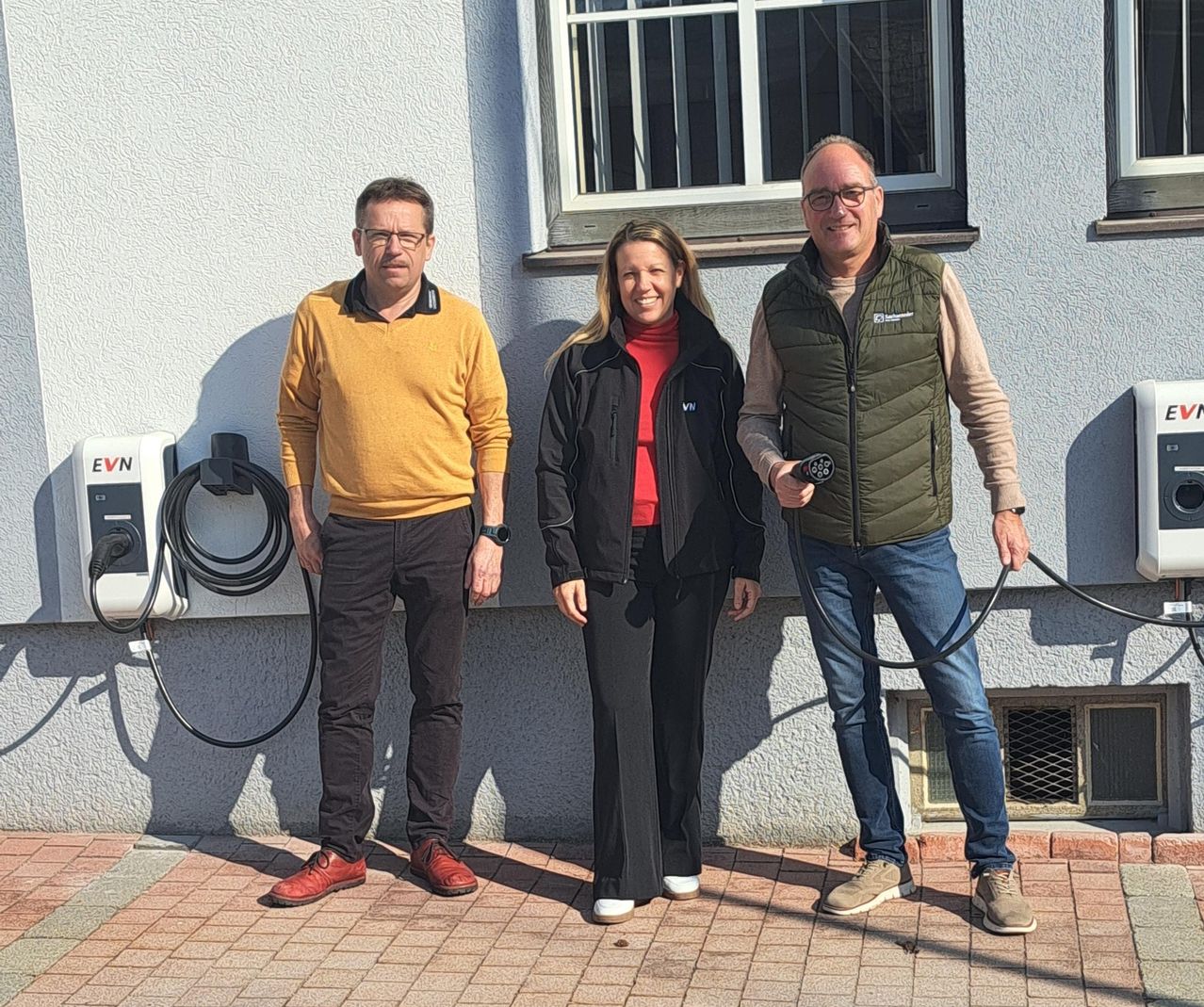 Three individuals stand next to a charging station in front of a white building. The man on the left wears a yellow sweater. The woman in the middle smiles with her hand on her hip. The man on the right holds a charging cable.