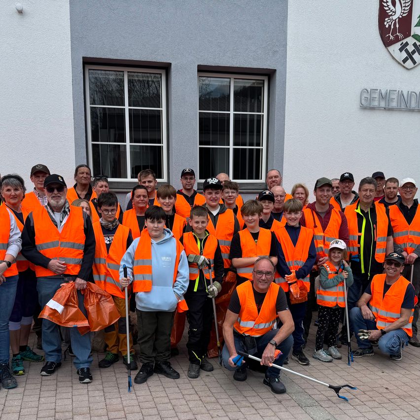 A group of people in orange safety vests pose for a photo outside a building, some holding tools. They appear to be participating in a community cleanup.