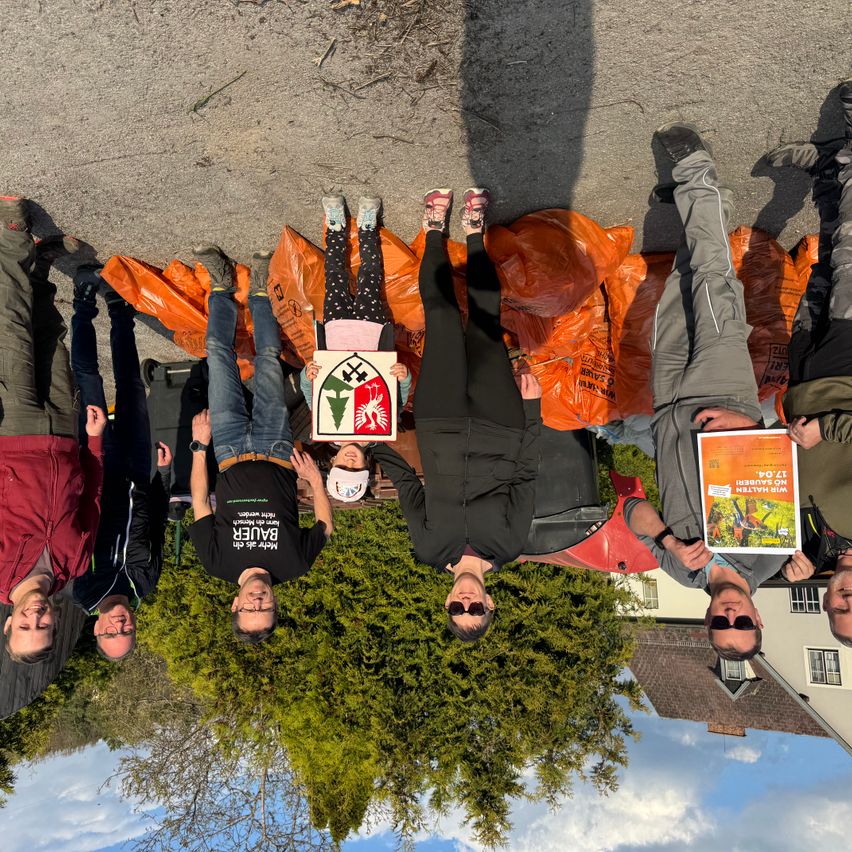 A group of people wearing jackets and pants, standing around a tree with a green sign, holding orange bags, and smiling for a photo.