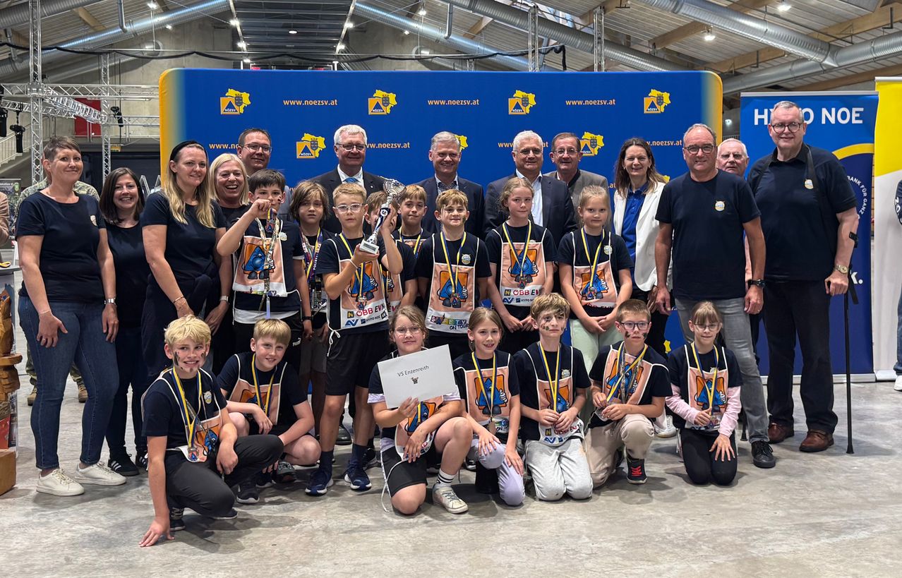 Eine Gruppe junger Teilnehmer und Erwachsene posiert für ein Foto vor einem Banner im Hintergrund. Die jungen Teilnehmer tragen Medaillen und halten einen Pokal und ein Zertifikat.