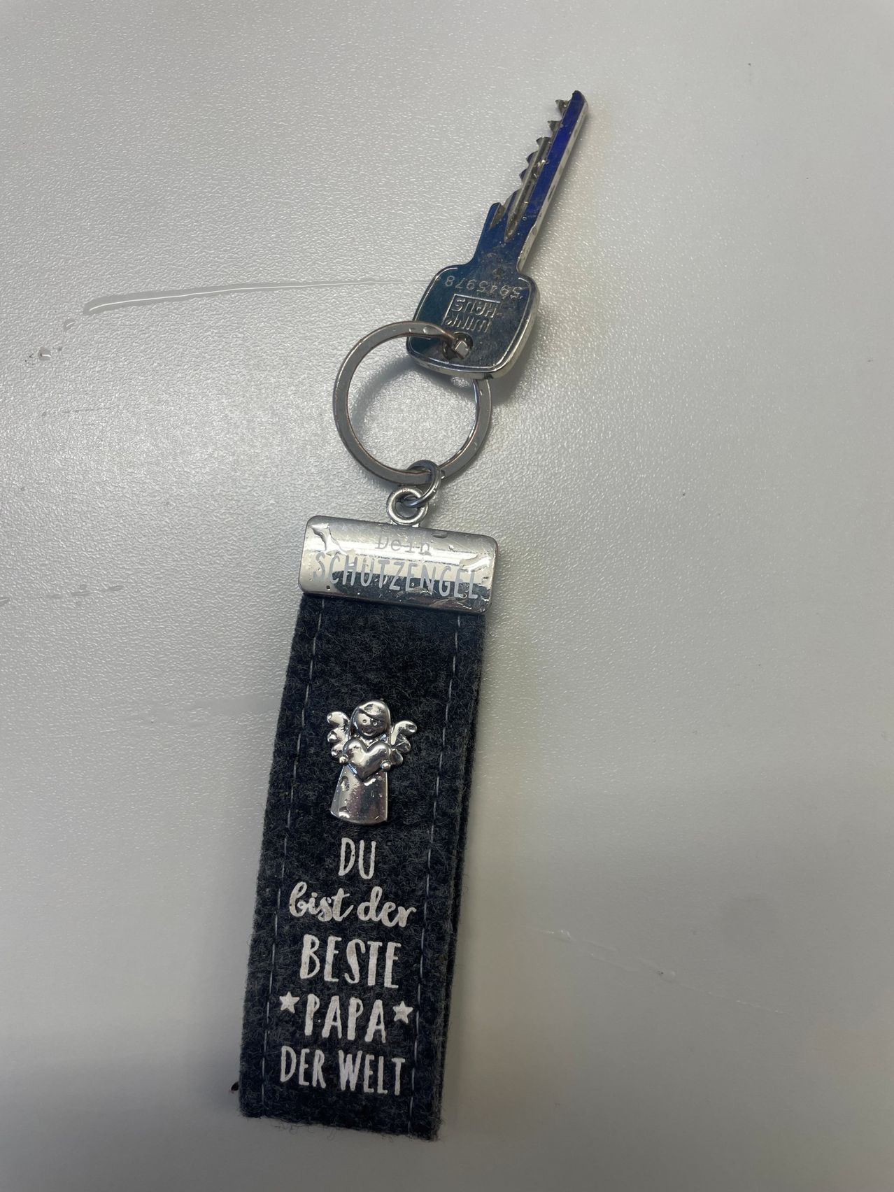 Ein silberner Schlüsselanhänger mit einem Schlüssel, einem schwarzen Band und einer kleinen Engelsfigur. Es steht 'Du bist der beste Papa' in silbernen Buchstaben.