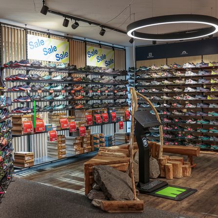 Ein Schuhgeschäft mit mehreren Regalen voller Sneaker im Angebot. Helle Schilder und kreisförmige Beleuchtung darüber. Holzbänke und ein Ausstellungsbereich mit Steinen.
