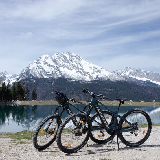 Zwei E-Bikes stehen auf Kies neben einem See mit verschneiten Bergen im Hintergrund.