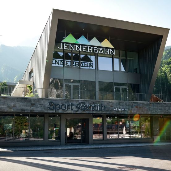 Das Jennerbahn Sport Resort zeigt ein modernes Gebäude mit großen Glasfenstern und einem Balkon vor einer Bergkulisse. Es enthält ein Schild für Sport Renthoth und Alpin Leben.