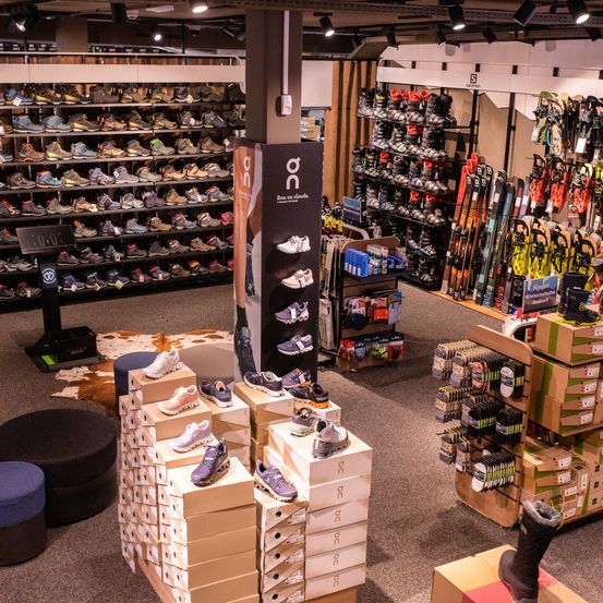 Ein Geschäft zeigt verschiedene Schuhe und Sportgeräte. Auf der linken Seite befinden sich mehrere Regale mit verschiedenen Schuhen, während auf der rechten Seite Skier und Snowboards zu sehen sind. In der Mitte befindet sich ein großes Schild mit einer Produktwerbung.