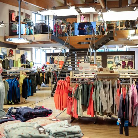 Ein Bekleidungsgeschäft gefüllt mit Kleiderständern voller farbenfroher Kleidung. Der Laden hat einen Holzboden und eine Treppe, die in die zweite Ebene führt. Es hängen viele Kleidungsstücke an den Stangen. Der Laden hat auch einen Monitor in der zweiten Ebene.