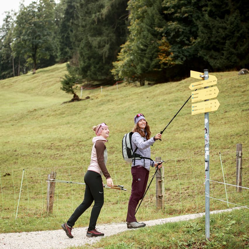 Zwei Frauen wandern in den Bergen. Sie lächeln und halten Trekkingstöcke. Ein gelber Schild mit Text und Zahlen ist an einem Pfosten befestigt.