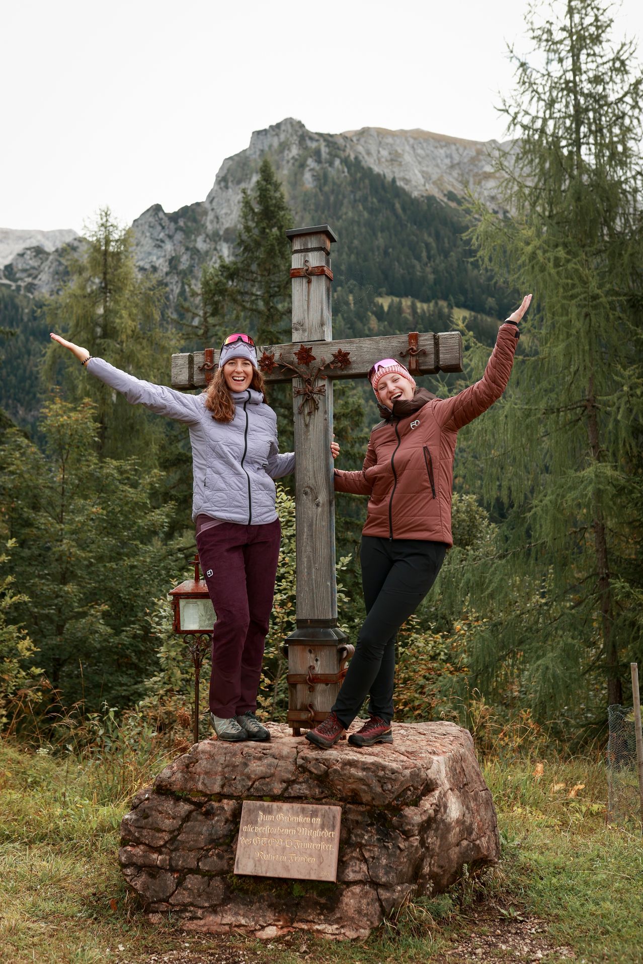 Zwei Frauen stehen auf einem Felsen und posieren für ein Foto mit einem Holzkreuz in einem bergigen, bewaldeten Gebiet. Sie tragen Winterkleidung.