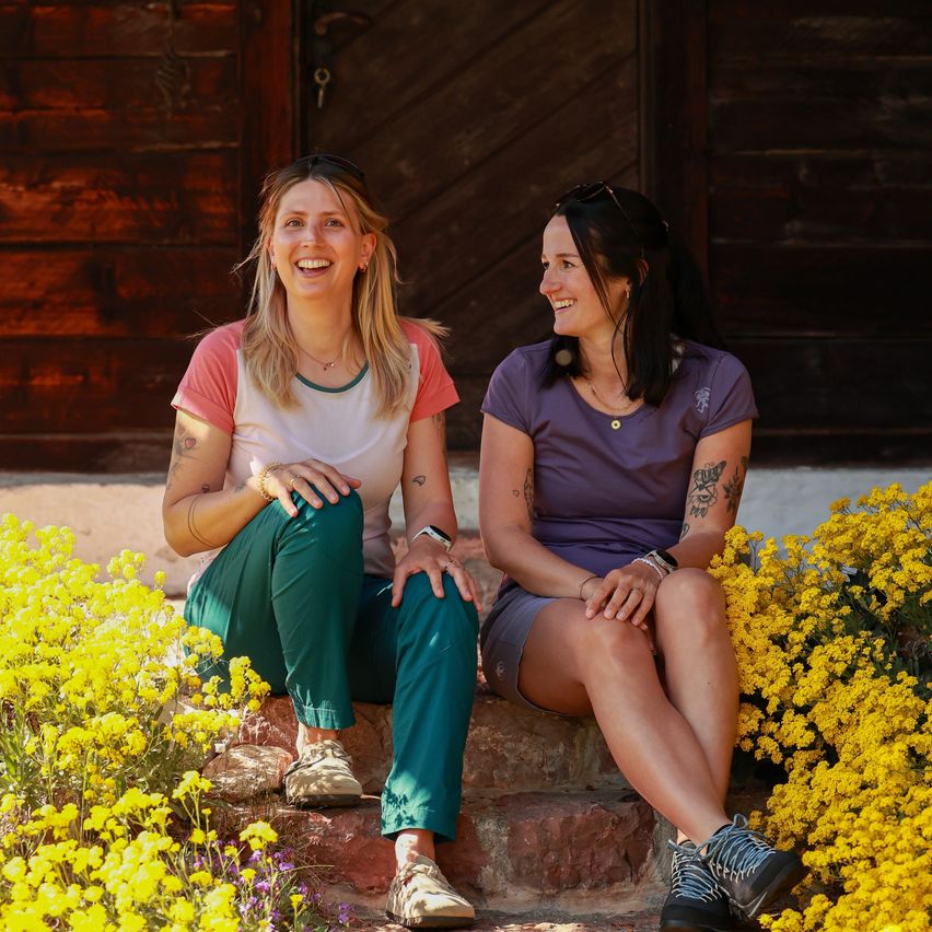 Two women sit on steps in front of a wooden building, surrounded by yellow flowers. They both wear necklaces, bracelets, and have tattoos. They smile for a photo.