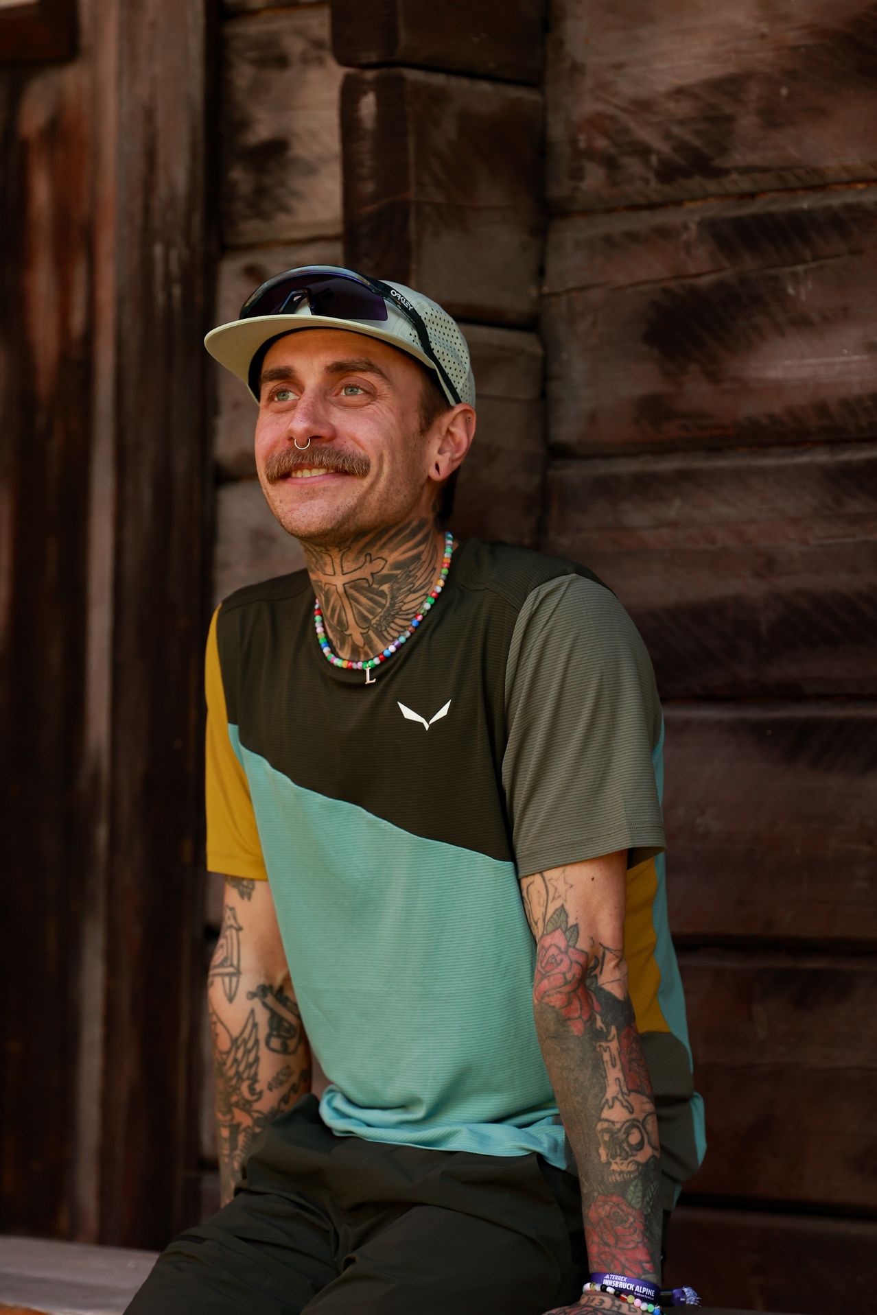 A man with tattoos and a colorful shirt is smiling and posing for a photo. He is wearing a hat with sunglasses on top.