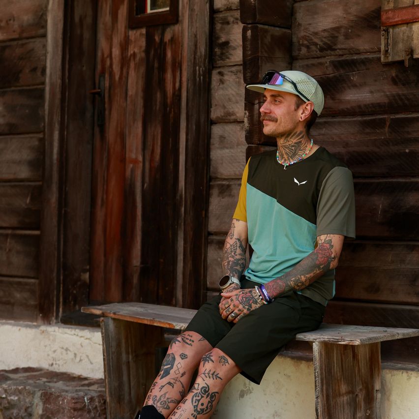 A man with tattoos on his arms and legs is sitting on a wooden bench outside a wooden house. He is wearing a cap, sunglasses, and a necklace.
