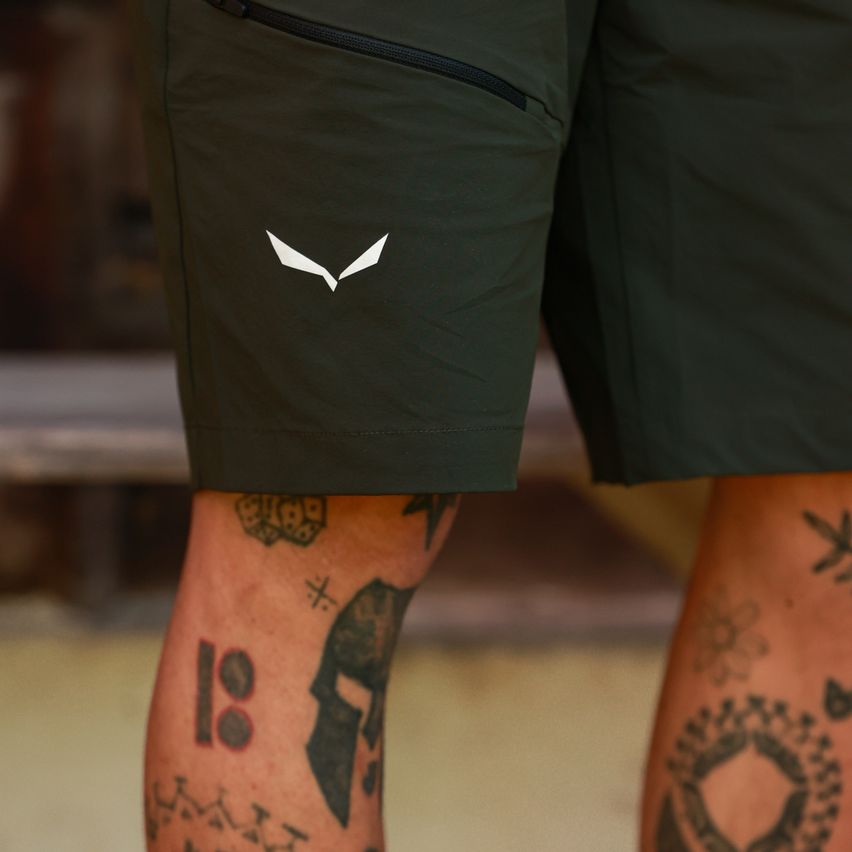 Close-up of a pair of olive green shorts with a white logo on the left leg. The person's tattooed legs are visible below the shorts.