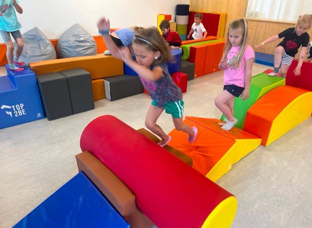 Mehrere Kinder spielen auf einer Matte in einem Spielzimmer. Ein Mädchen springt über eine rote Matte und ein anderes Mädchen läuft auf der Matte.