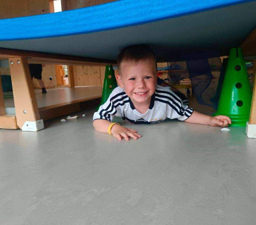 Ein junger Junge liegt lächelnd mit den Händen auf dem Boden auf einem grauen Boden unter einem blauen Tisch. Grüne Kegel sind um ihn herum aufgestellt. Eine Person steht hinter ihm.