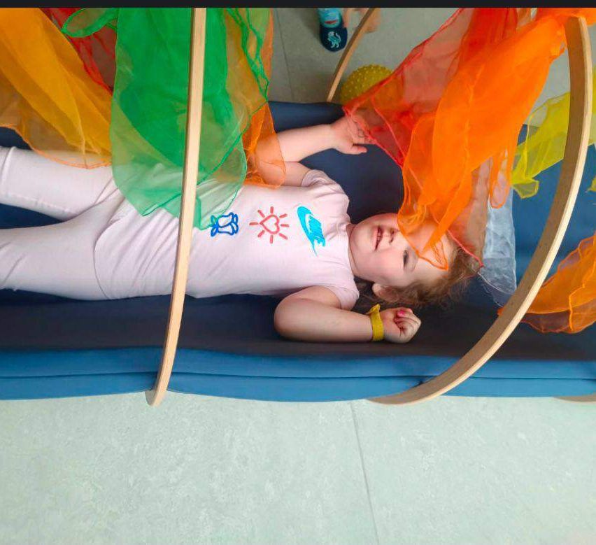 Ein Kleinkind liegt auf einer blauen Oberfläche, umgeben von farbenfrohen, fließenden Stoffen. Sie trägt ein rosa Shirt mit verspielten Designs und ein gelbes Armband.