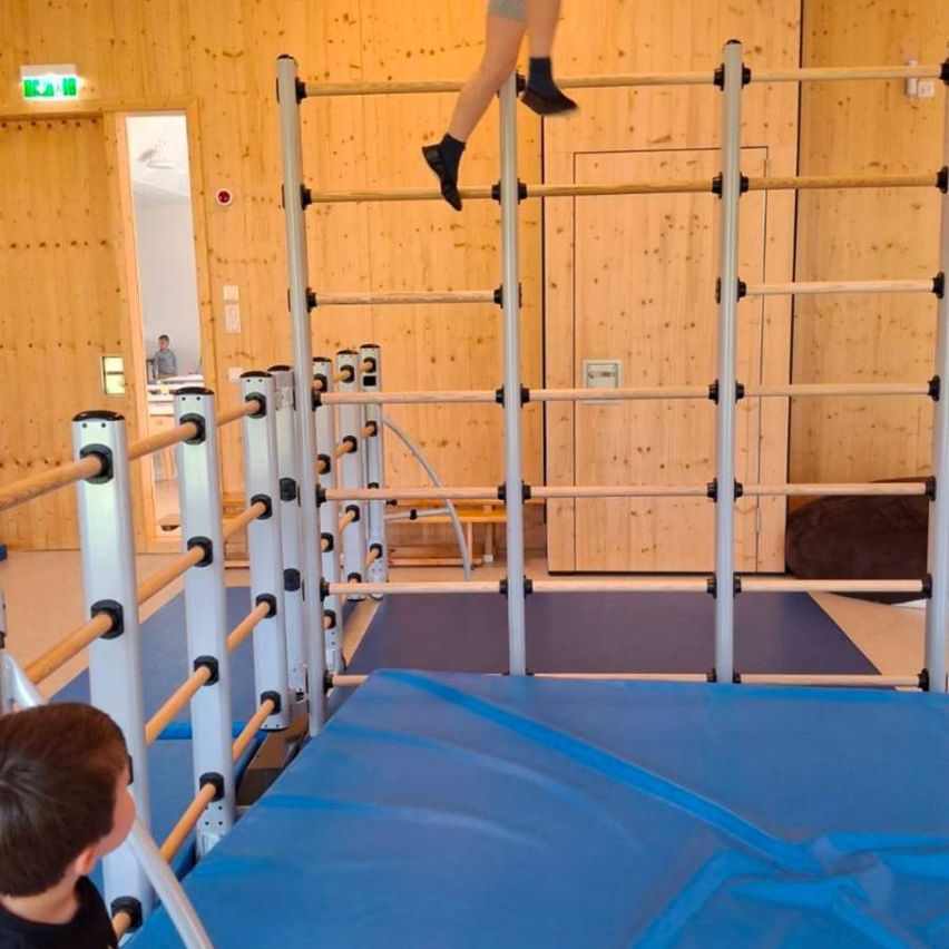 Ein junger Junge beobachtet ein anderes Kind, das eine Reihe von Parallelbarren in einem Fitnessstudio erklimmt. Das Fitnessstudio hat Holzwände und eine blaue Matte auf dem Boden.