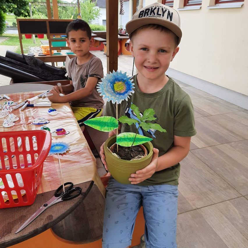 Zwei Jungen basteln Blumen im Klassenzimmer, einer hält ein blumenähnliches Objekt und der andere sitzt an einem Schreibtisch mit verschiedenen Bastelmaterialien.