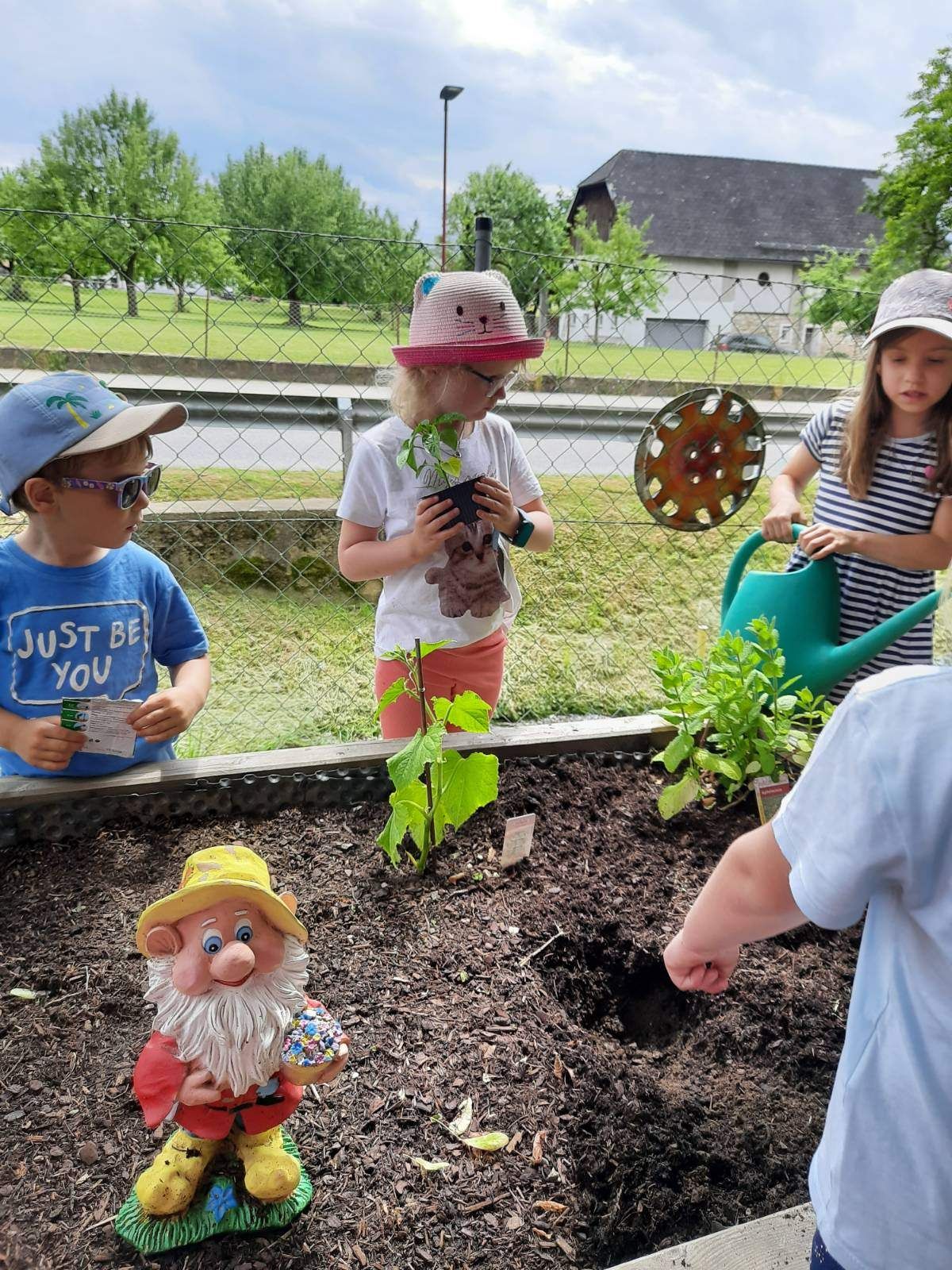 Kinder pflanzen Blumen in einem Garten mit einem Zaun, einem Haus und einem Auto im Hintergrund. Ein Kind gießt Pflanzen.