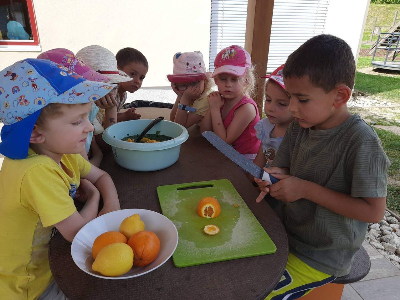 Kinder in Hüten sitzen um einen Tisch herum, ein Junge schneidet eine Orange. Schüsseln mit Zitronen und Orangen stehen auf dem Tisch. Ein anderes Kind schaut zu.
