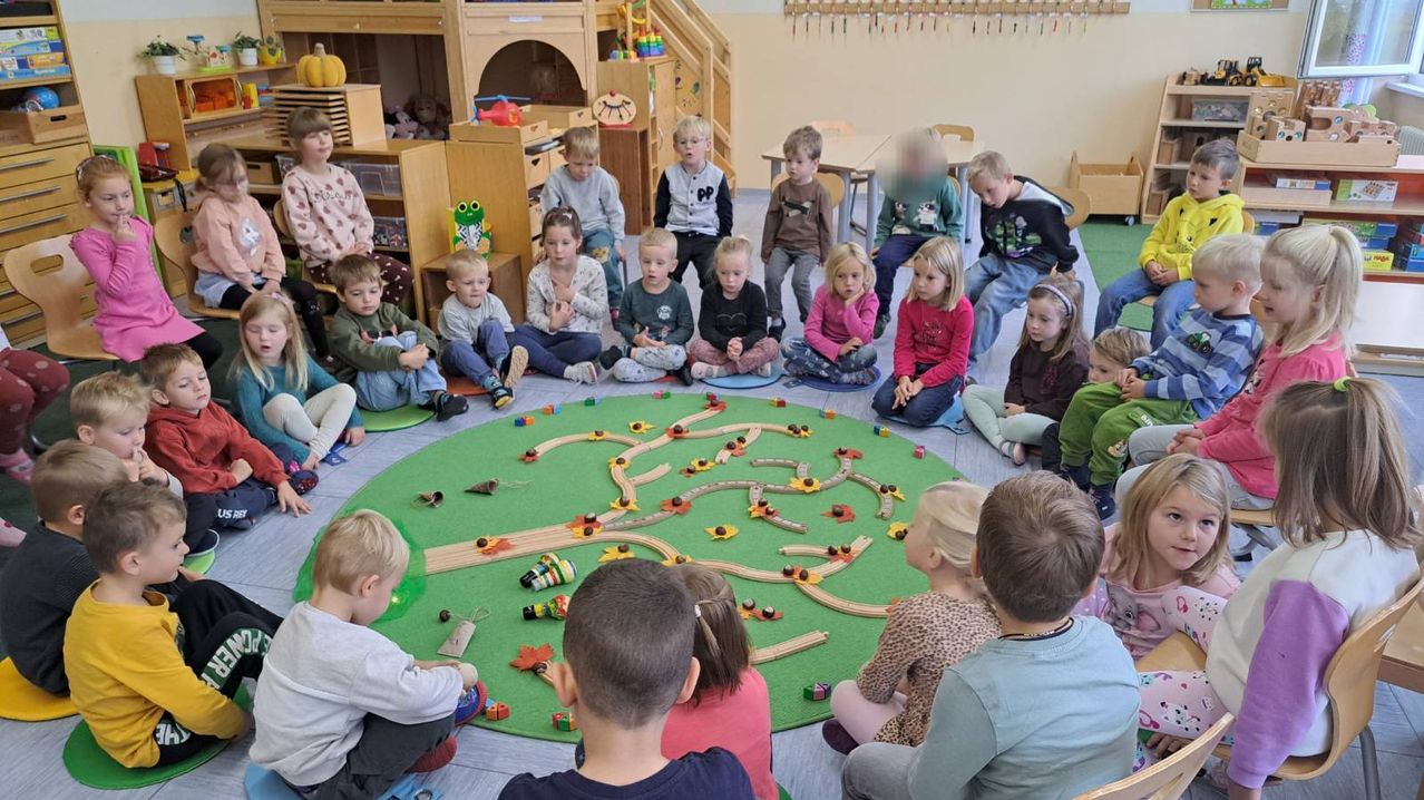 Eine Gruppe von Kindern ist in einem Kreis um einen grünen Teppich mit einem Spielzeug-Zug-Set versammelt. Einige sitzen auf dem Boden, andere auf Kissen. Sie scheinen an einer Geschichte oder Aktivität beteiligt zu sein.