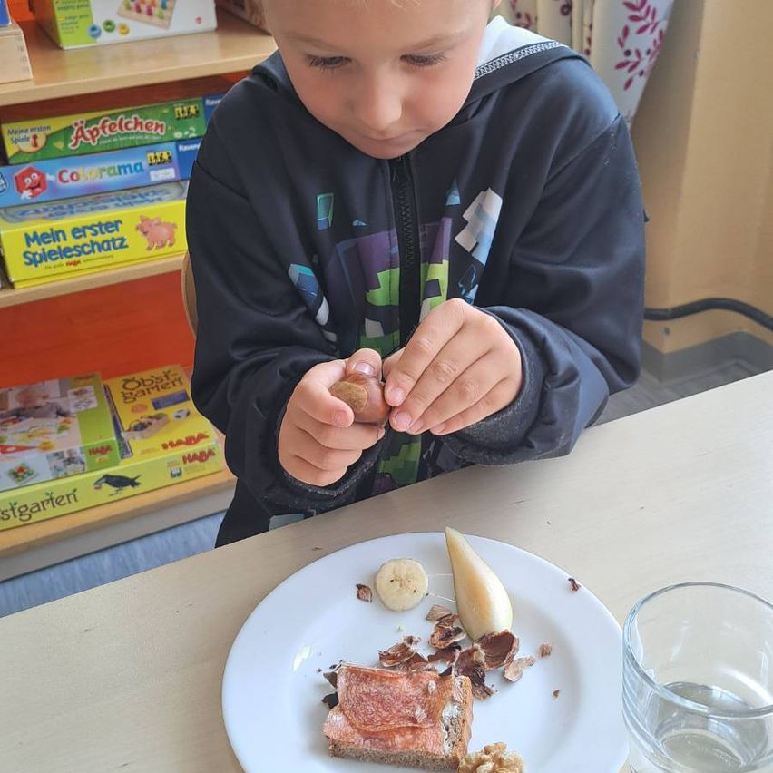 Ein junger Junge isst ein Sandwich und Nüsse, während er an einem Tisch sitzt. Vor ihm steht ein Teller mit Essen und ein Glas Wasser. Hinter ihm befindet sich ein Regal mit Kinderbüchern und Spielzeug.