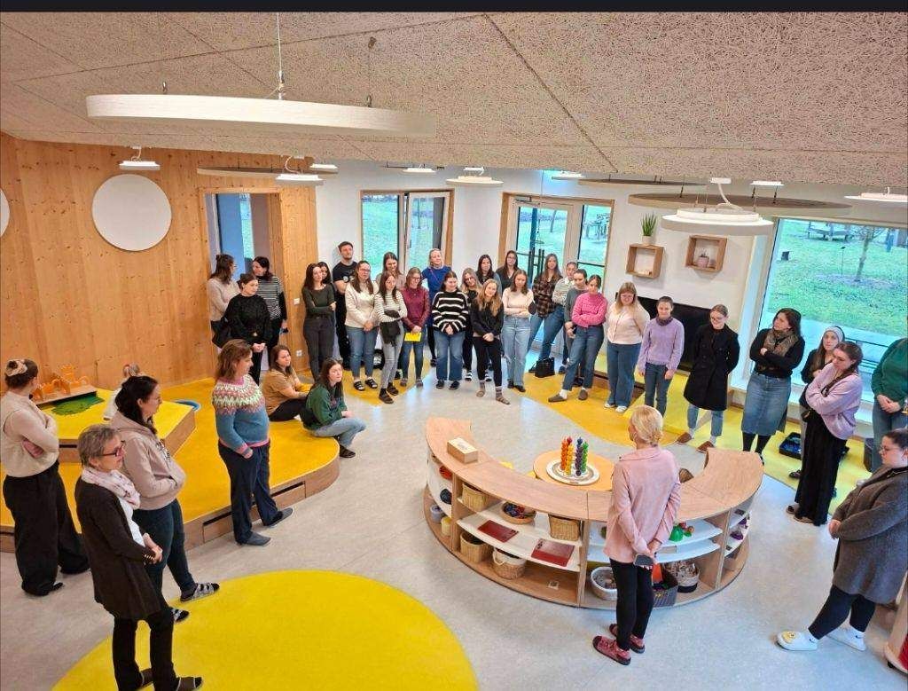 Eine Gruppe von Menschen versammelt sich in einem modernen Raum mit Holzboden, einem Holztisch und Glastüren. Einige stehen, andere sitzen auf gelben Plattformen.