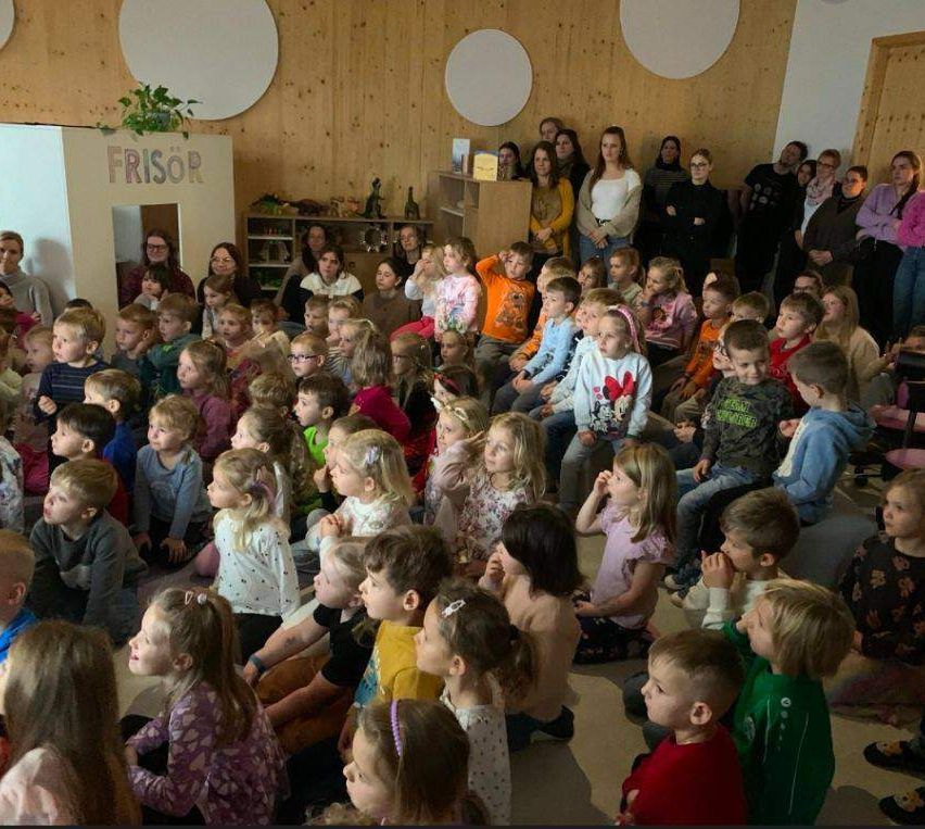 Eine große Gruppe von Kindern und Erwachsenen versammelt sich in einem Raum mit Holzwänden. Die Kinder sitzen im Kreis, während die Erwachsenen hinter ihnen stehen. Ein Schild mit der Aufschrift Frisör ist an der Wand angebracht.