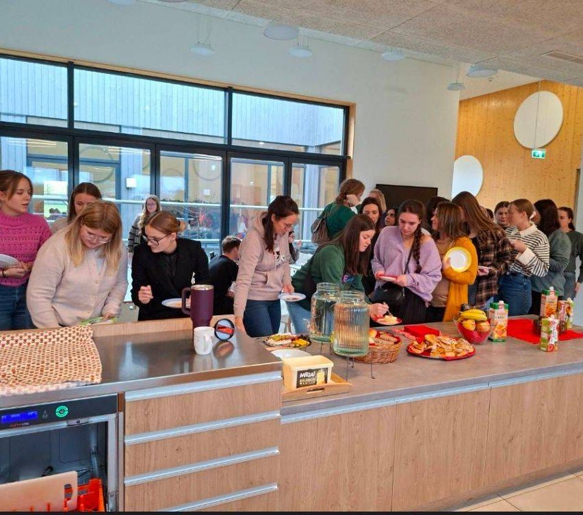 Eine Gruppe von Frauen in einem Raum mit einem Buffettisch voller Essen, Getränke und Teller. Einige Frauen stehen, während andere sitzen, und sie essen alle. Der Raum hat Glasfenster und eine Holzwand.