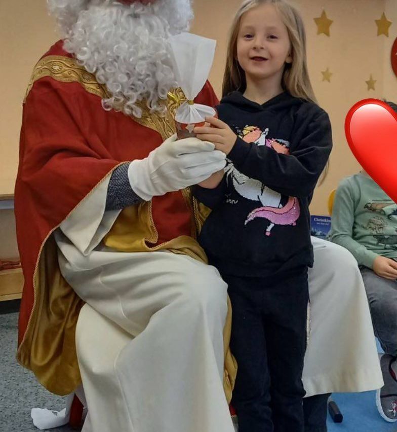 Ein fröhliches Mädchen mit einem Einhorn-Shirt steht neben einer als Weihnachtsmann verkleideten Person, die ein Geschenk hält. Ein weiteres Kind sitzt im Hintergrund.