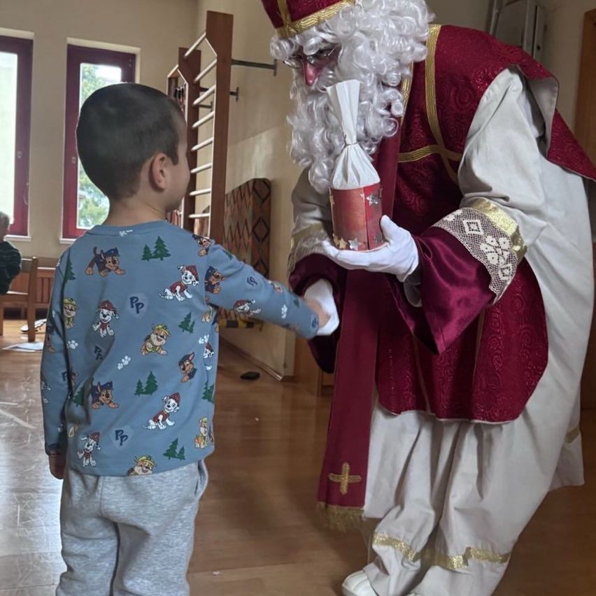 Ein junger Junge in Pyjamas erhält von einer als Weihnachtsmann verkleideten Person ein Geschenk in einem Raum mit Holzboden und Fenstern.