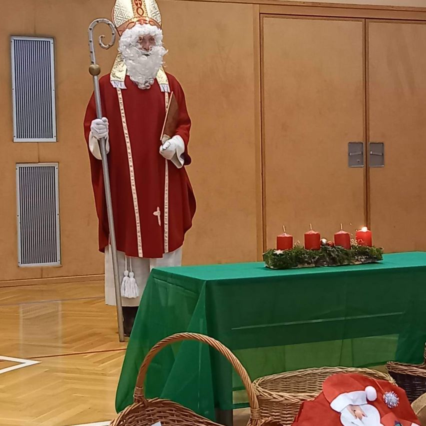 Eine Person in St.-Nikolaus-Verkleidung steht vor einem Tisch mit grünem Tuch und einer Kerzengirlande. Ein Weihnachtshut liegt auf einem der Körbe auf dem Boden.