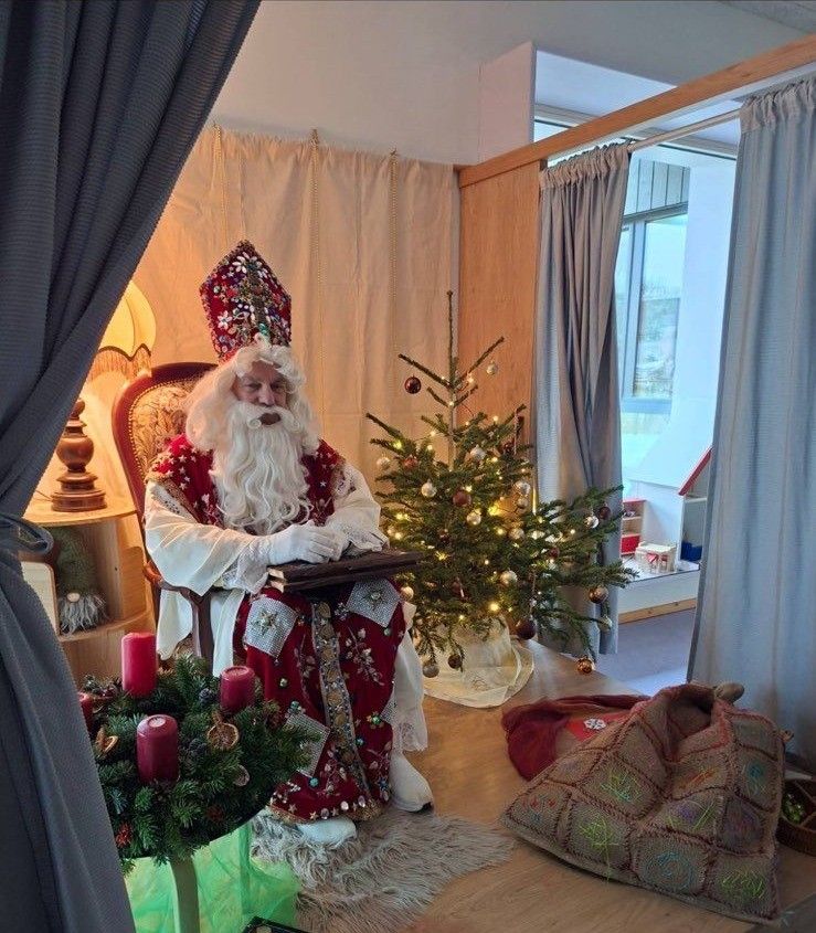 Ein älterer Mann, der als Weihnachtsmann verkleidet ist, sitzt auf einem Stuhl vor einem geschmückten Weihnachtsbaum. Es gibt rote Kerzen, eine Lampe und einen kleinen Weihnachtsbaum auf dem Boden.
