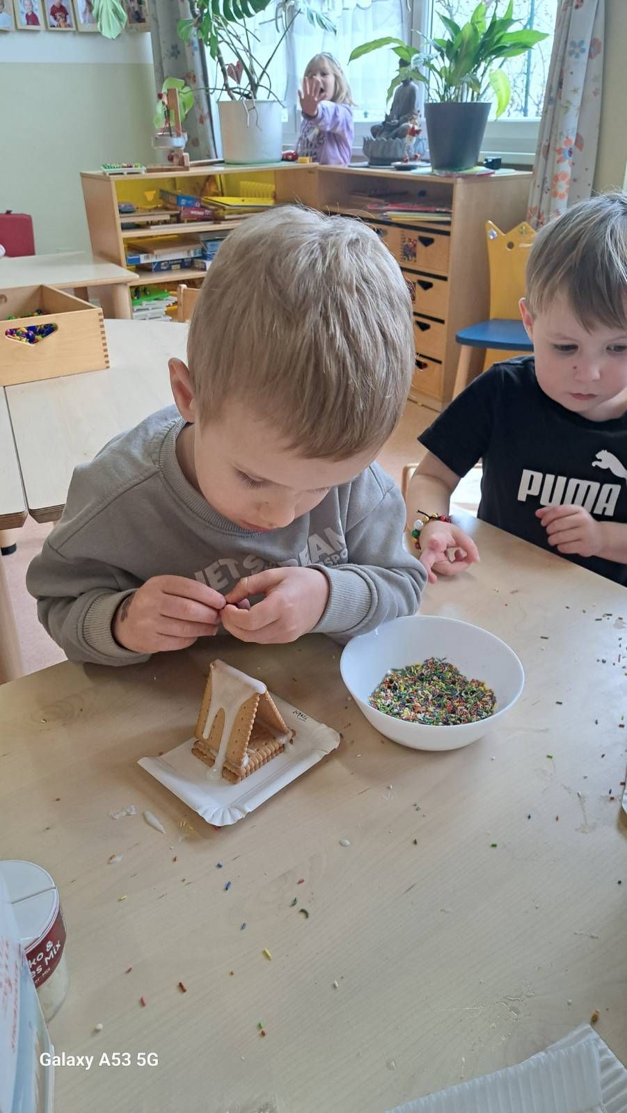 Zwei junge Jungen sind bei einer Bastelaktivität beschäftigt und dekorieren ein Lebkuchenhaus mit bunten Streuseln. Ein Junge platziert sorgfältig die Streusel auf dem Haus, während der andere zusieht.