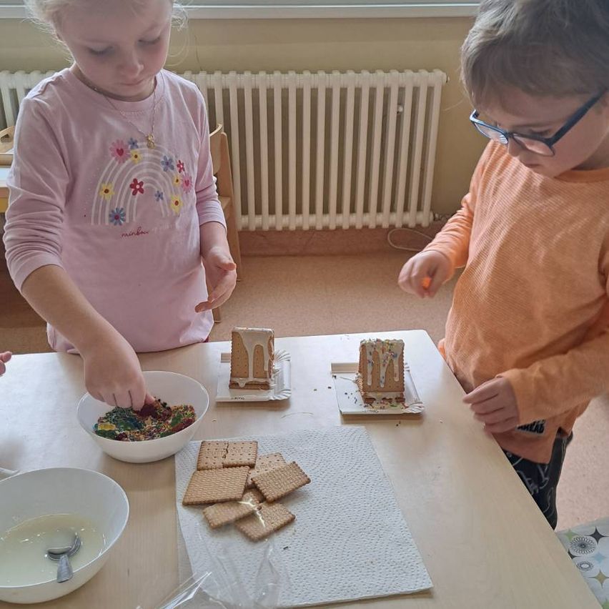 Zwei Kinder dekorieren Lebkuchenhäuser mit Zuckerguss und Streuseln an einem Tisch. Das Mädchen trägt ein rosa T-Shirt mit Regenbogen-Design, und der Junge trägt eine Brille.