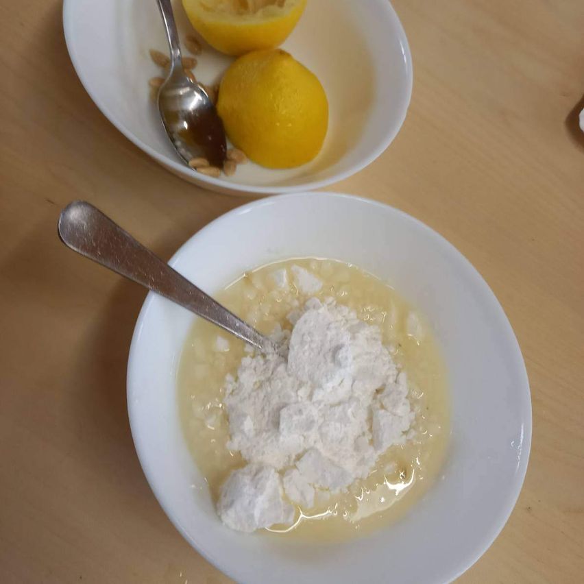 Zwei weiße Schüsseln auf einem Holztisch, eine mit Mehl und Flüssigkeit, die andere mit Zitronen und einem Löffel.
