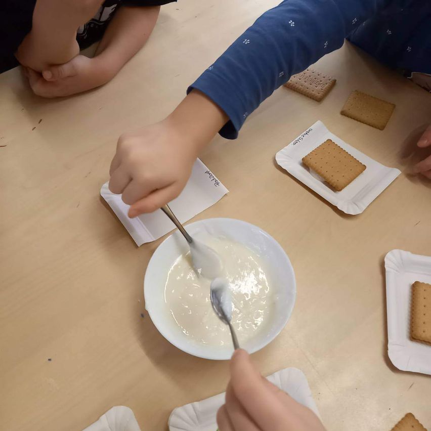 Zwei Kinder sitzen an einem Holztisch und sind mit einer Aktivität beschäftigt. Ein Kind verwendet einen Löffel, um etwas in einer weißen Schüssel zu mischen. Mehrere Cracker sind auf weißen Servietten auf dem Tisch platziert.