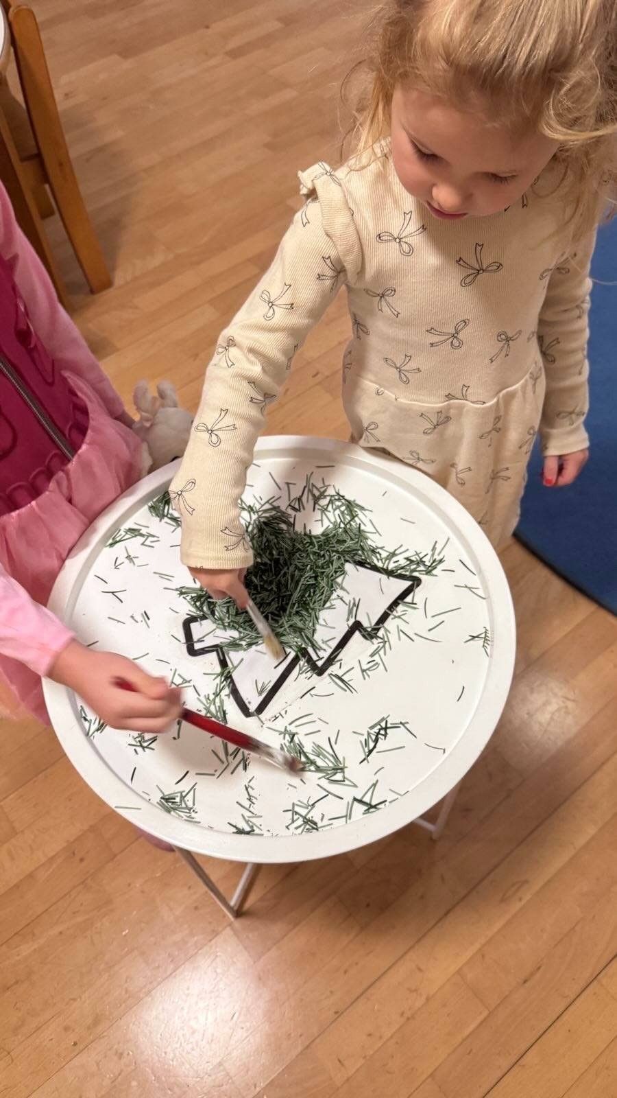 Zwei Kinder stehen neben einem Tisch mit einem Weihnachtsbaumdesign darauf. Sie verwenden Pinsel, um grünen Gras über das Design zu verteilen.