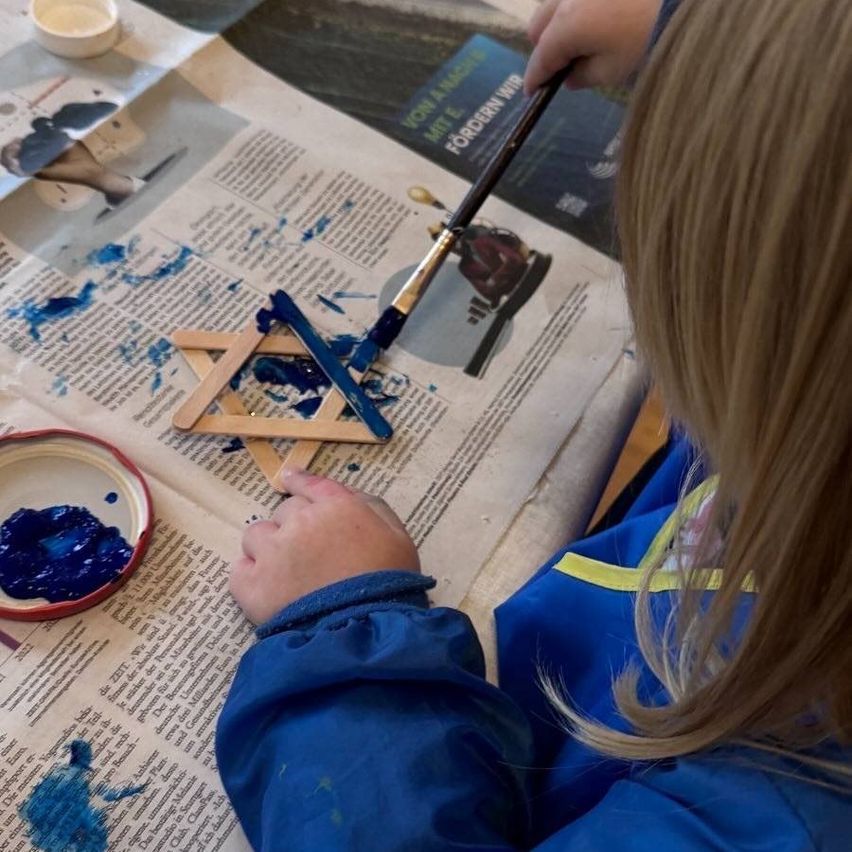 Ein Kind malt einen Davidstern mit blauer Farbe auf Zeitungspapier. Ein Farbtopf und ein Pinsel liegen in der Nähe.