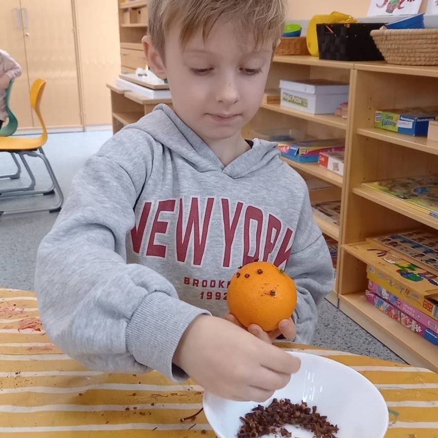 Ein junger Junge schält eine Orange, während er an einem Tisch im Klassenzimmer sitzt. Er trägt eine graue Hoodie mit 'NEW YORK' darauf.