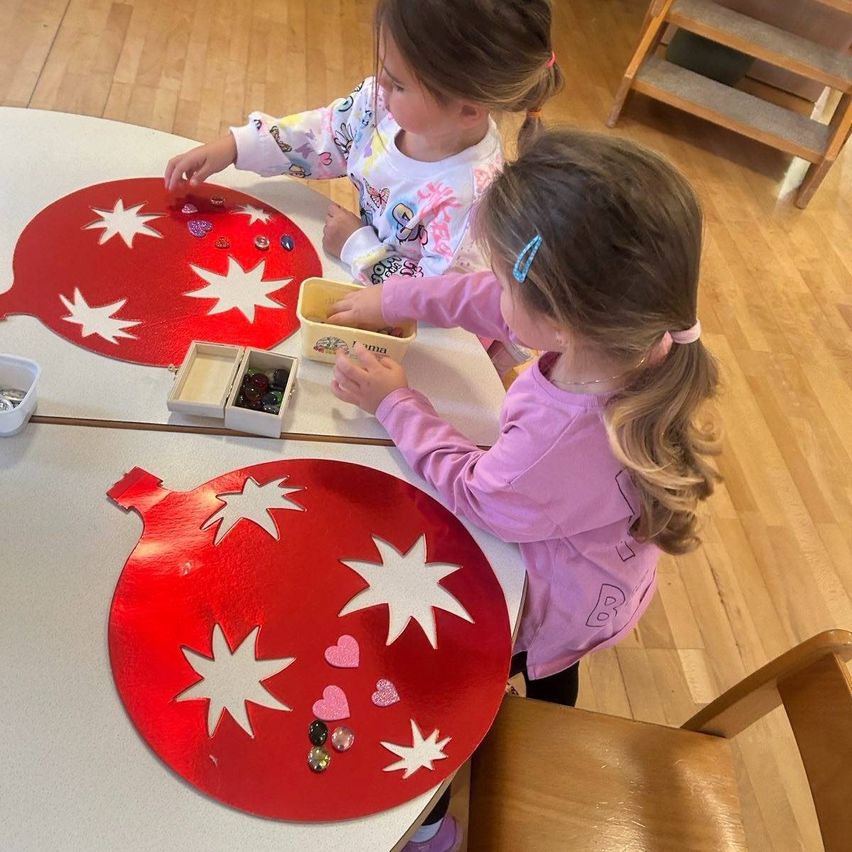 Zwei kleine Mädchen schmücken einen Weihnachtsschmuck am Tisch, eine platziert Perlen und die andere hält einen Behälter. Der Schmuck ist rot mit weißen Sternmustern.