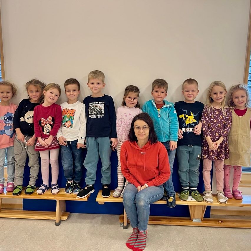 Eine Gruppe von Kindern, die in zwei Reihen auf Bänken stehen, mit einer Frau, die auf einer der Bänke vor ihnen sitzt. Sie lächeln und scheinen für ein Foto zu posieren.