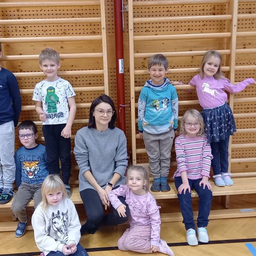 Eine Gruppe von Kindern und einer erwachsenen Frau stehen und sitzen in einer Turnhalle. Die Frau kniet vor den Kindern. Sie tragen alle Winterkleidung. Der Hintergrund ist eine Holzwand.