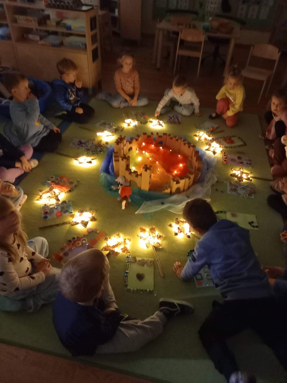 Kinder sitzen im Kreis um eine beleuchtete Burgdekoration. Jedes Kind hat ein kleines Licht. Sie sind in einer Aktivität verstrickt, möglicherweise ein Geschichtenerzählen oder Basteln.