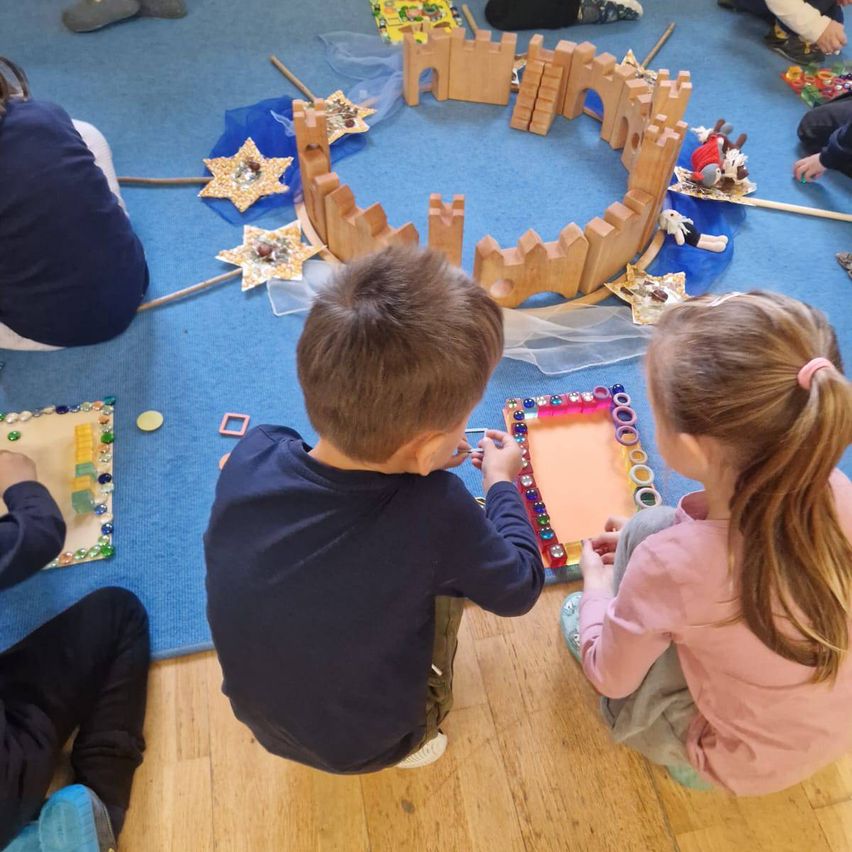 Eine Gruppe von Kindern spielt mit Holzklötzen und anderen Spielzeugen auf einem blauen Teppich. Ein Junge und ein Mädchen sitzen, während andere stehen.