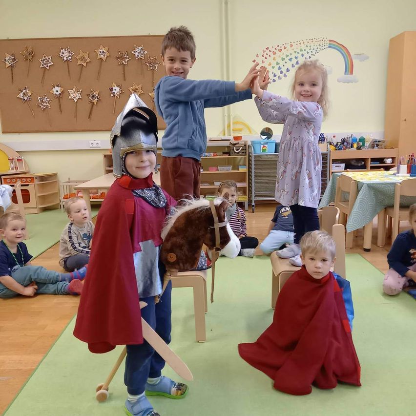 In einem Klassenzimmer sind eine Gruppe von Kindern in Kostümen gekleidet. Ein Junge mit Helm und Schwert steht mit einem Mädchen, während andere auf Stühlen oder dem Boden sitzen.