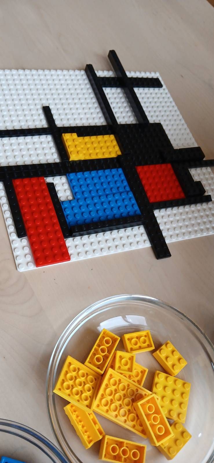 Ein farbenfrohes abstraktes Design aus LEGO-Steinen mit roten, blauen, gelben und schwarzen Teilen, angeordnet auf einer weißen Grundplatte. In der Nähe befindet sich eine Glasschale mit gelben LEGO-Steinen.