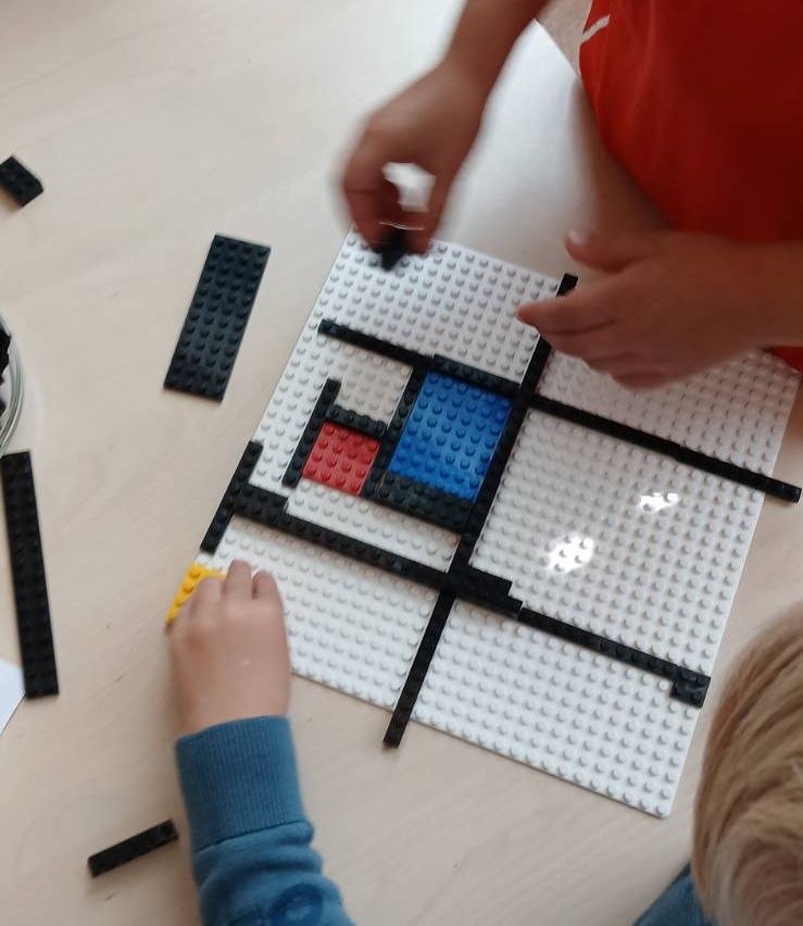 Zwei Kinder bauen ein Quadrat mit weißen, blauen, roten und schwarzen Lego-Steinen. Das Kind in Blau hält ein gelbes Stück.