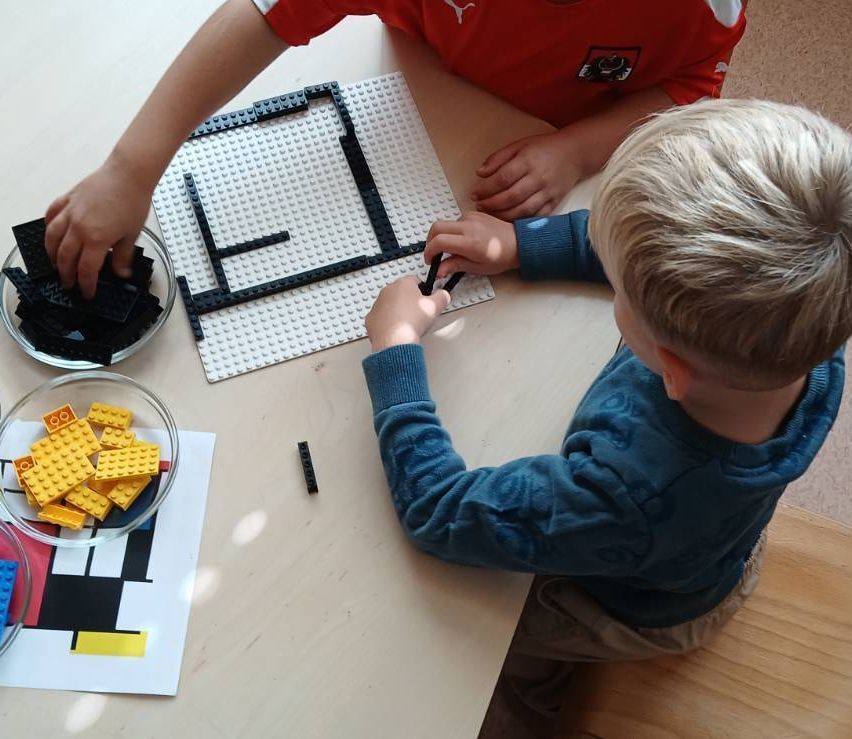 Zwei Kinder spielen mit Legos auf einem weißen Tisch. Ein Kind baut eine Struktur, während das andere zuschaut. Eine Schüssel mit Legos steht in der Nähe.